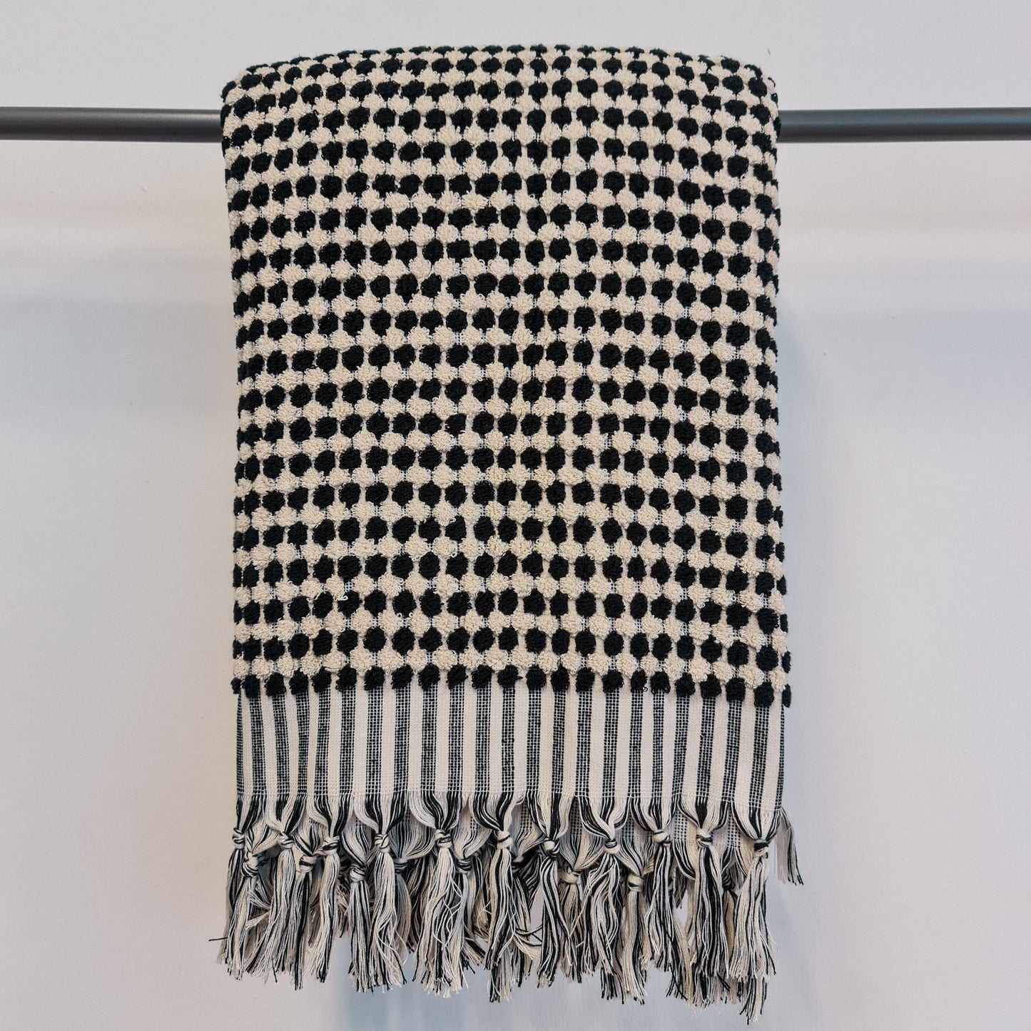 A black and white dotted Turkish hammam bath towel with fringes displayed on a hanger against a white background.