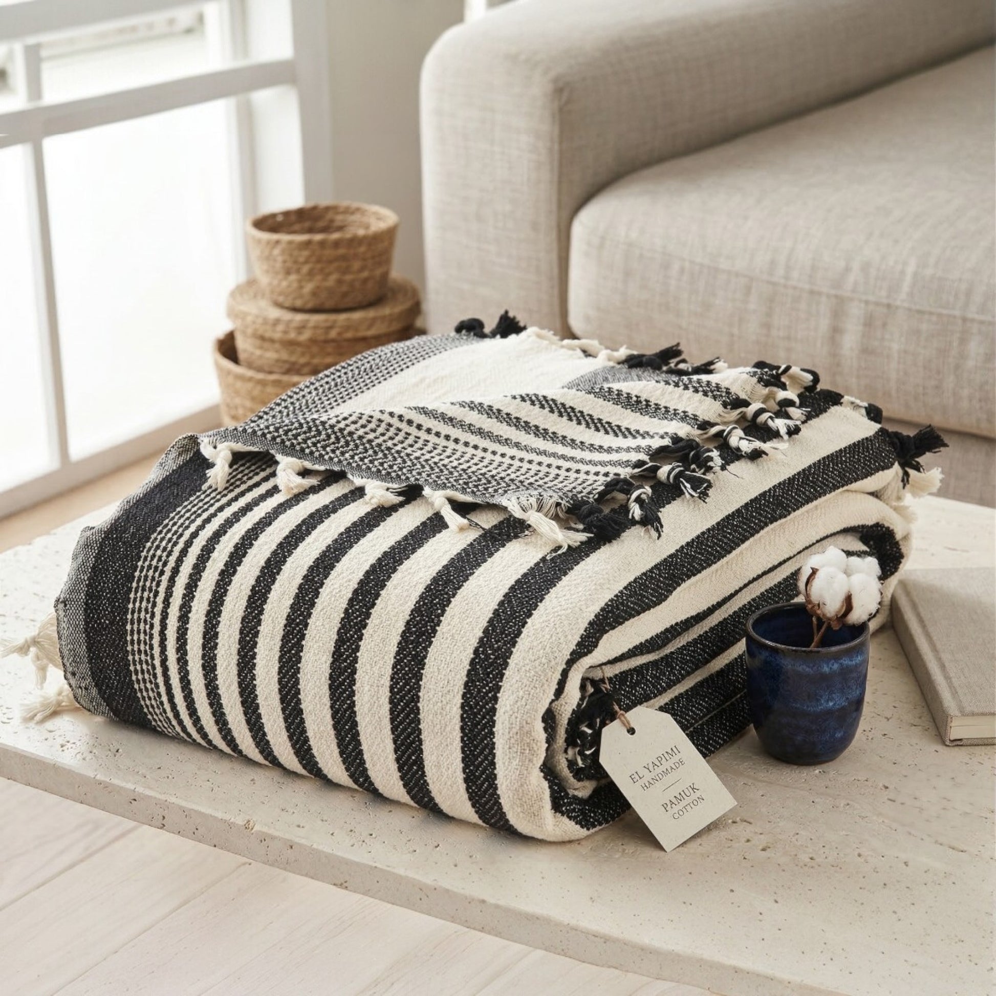 Black and white striped blanket with tassels on a wooden surface, with a neutral-colored sofa and woven baskets in the background.