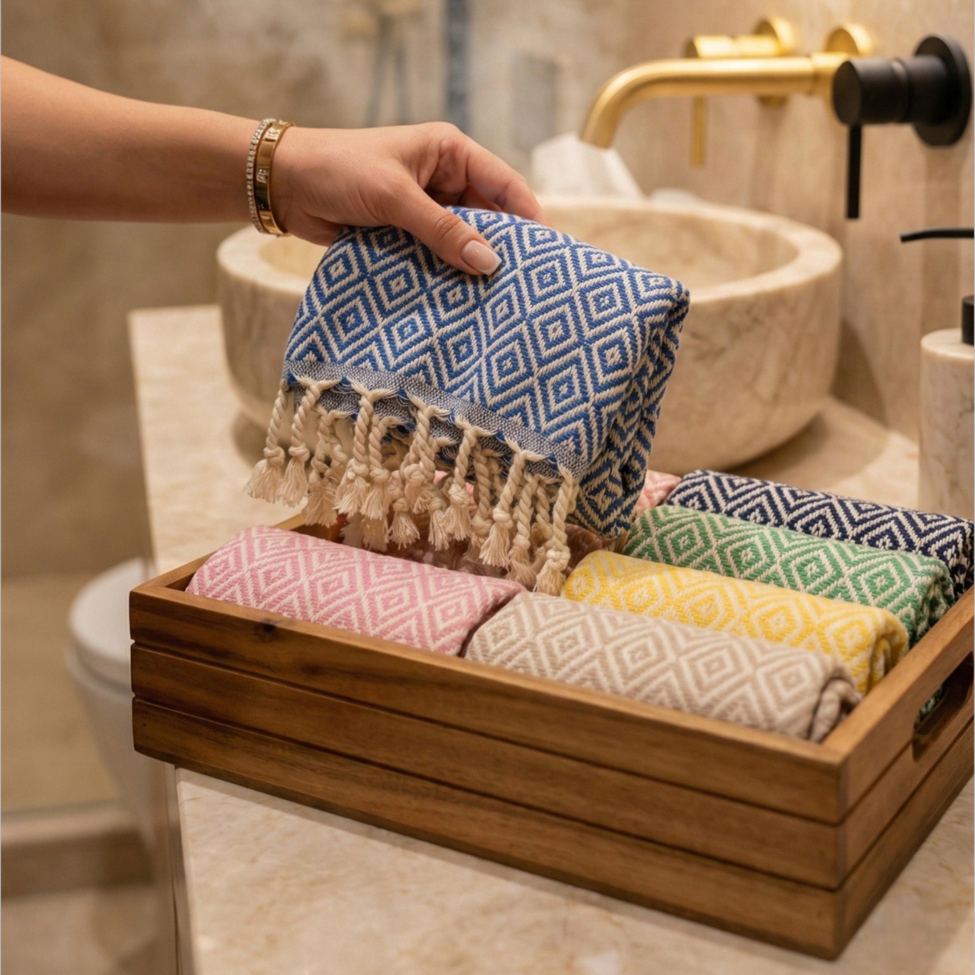 Hand holding a blue patterned Diamond Design Turkish Hand Towel  Handwoven Natural Cotton Sustainable Anatolian Craft over a wooden tray with colorful towels in a bathroom setting.