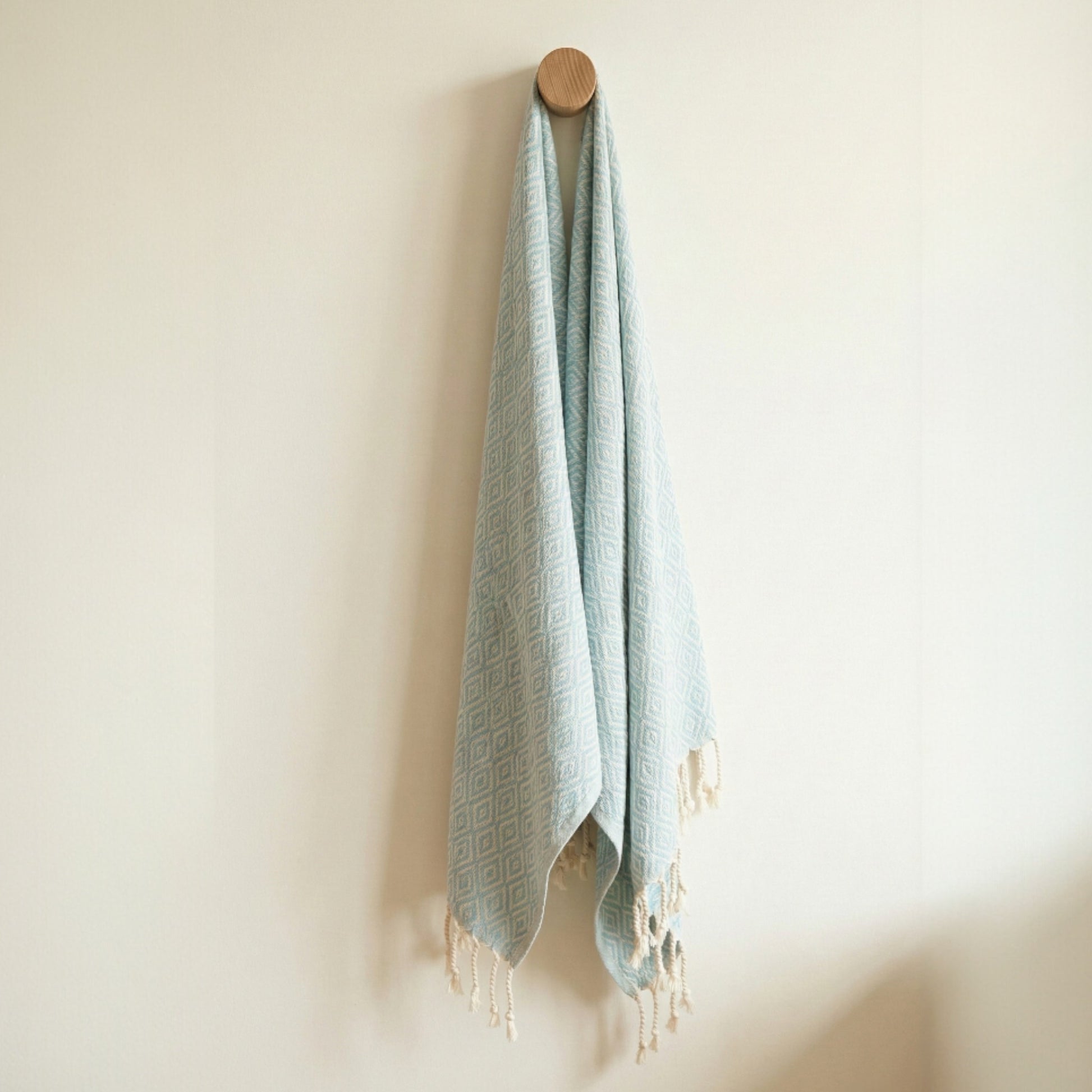 Light blue Diamond Design Turkish Hand Towel  Handwoven Natural Cotton Sustainable Anatolian Craft with tassels hanging on a wooden hook against a beige wall.