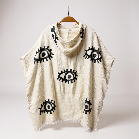 Beige poncho with black eye patterns on a wooden hanger against a white background