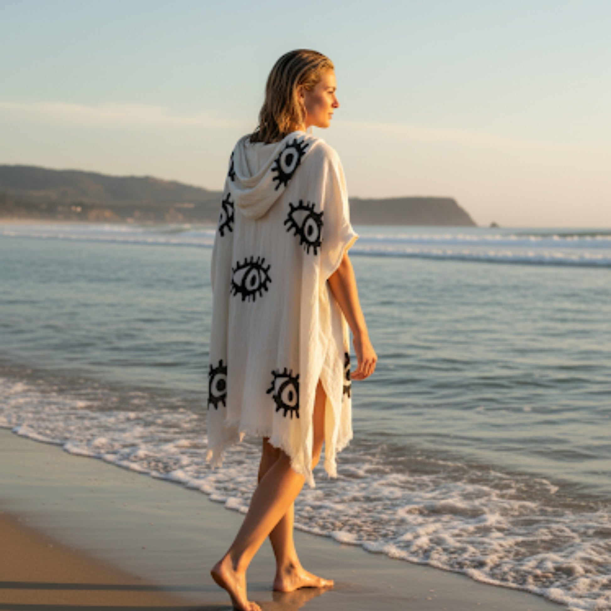 A person wearing a unisex, one-size-fits-all Turkish towel surf poncho. The poncho is hand-woven from natural ecru cotton and features intricate, hand-printed black Evil Eye patterns, printed in natural dyes. The poncho is versatile, used as a cover-up or a dress.