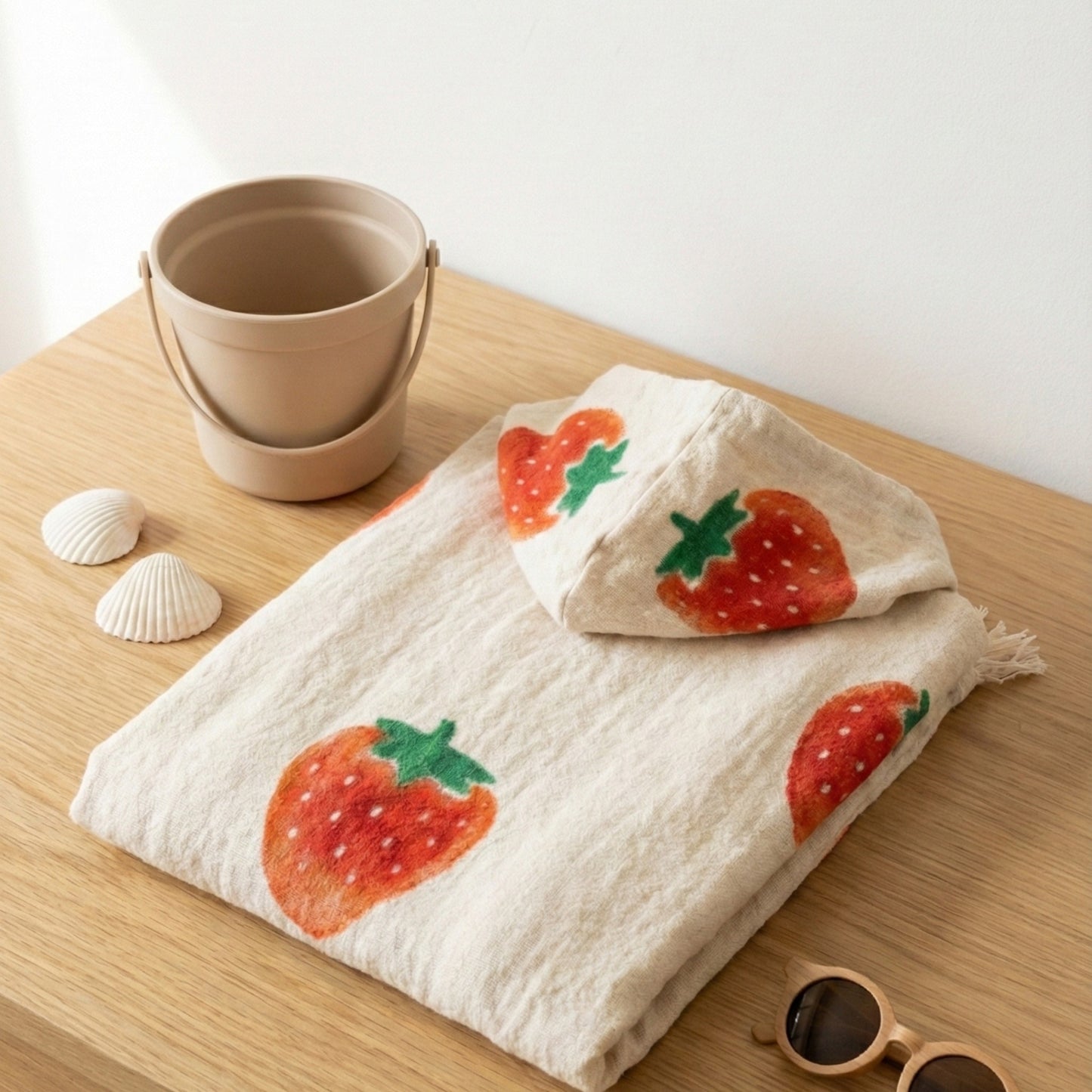 Folded Handmade Natural Cotton Kids Poncho – Strawberry Design on a wooden surface with a bucket and shells.