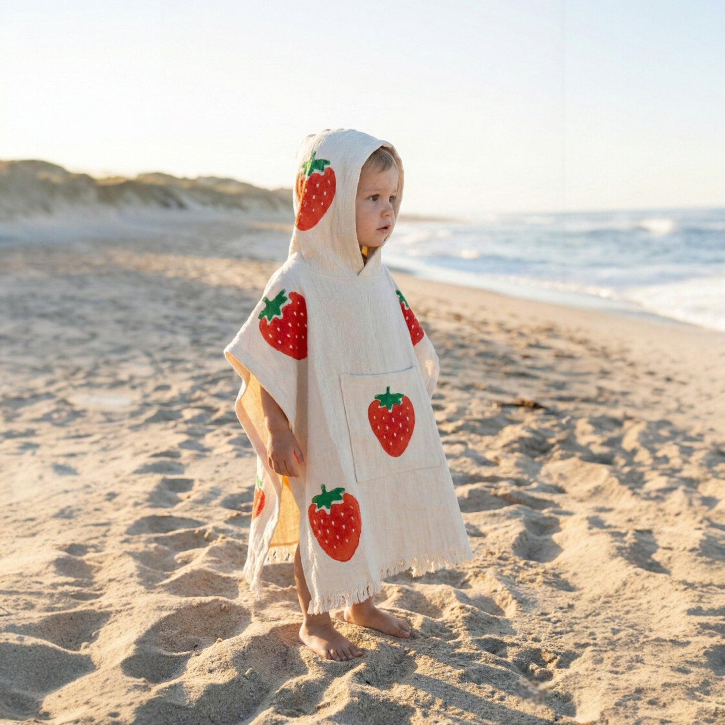 Child wearing a Handmade Natural Cotton Kids Poncho – Strawberry Design on a sandy beach.