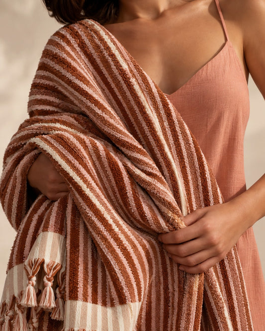 Woman wrapped in a handwoven Turkish terry bath towel made from 100% long-staple Turkish cotton, featuring brown and dusty pink stripes, plush absorbent texture, and hand-knotted tassels.