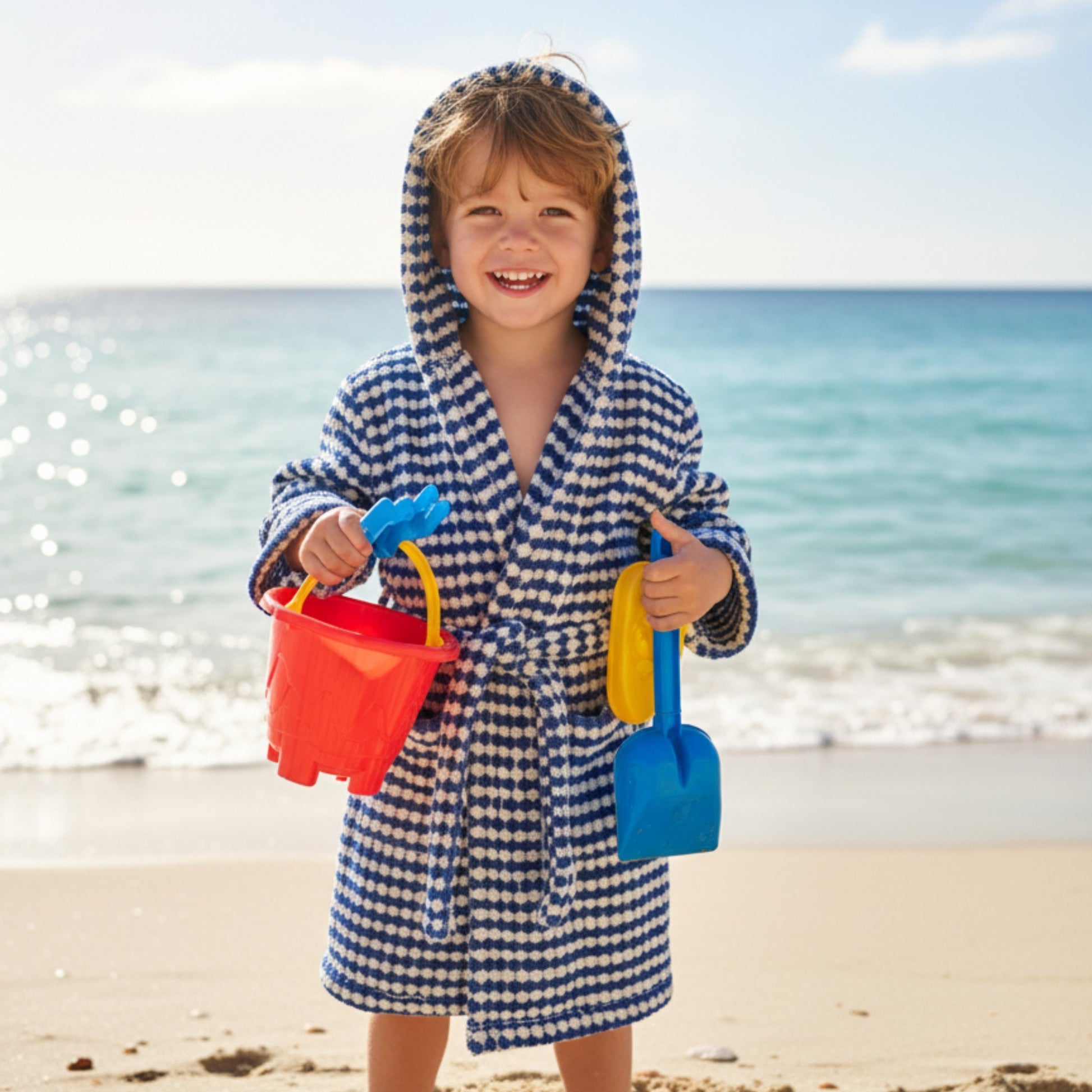 Child in a blue and white checkered robe holding beach toys on a sandy beach with ocean in the background