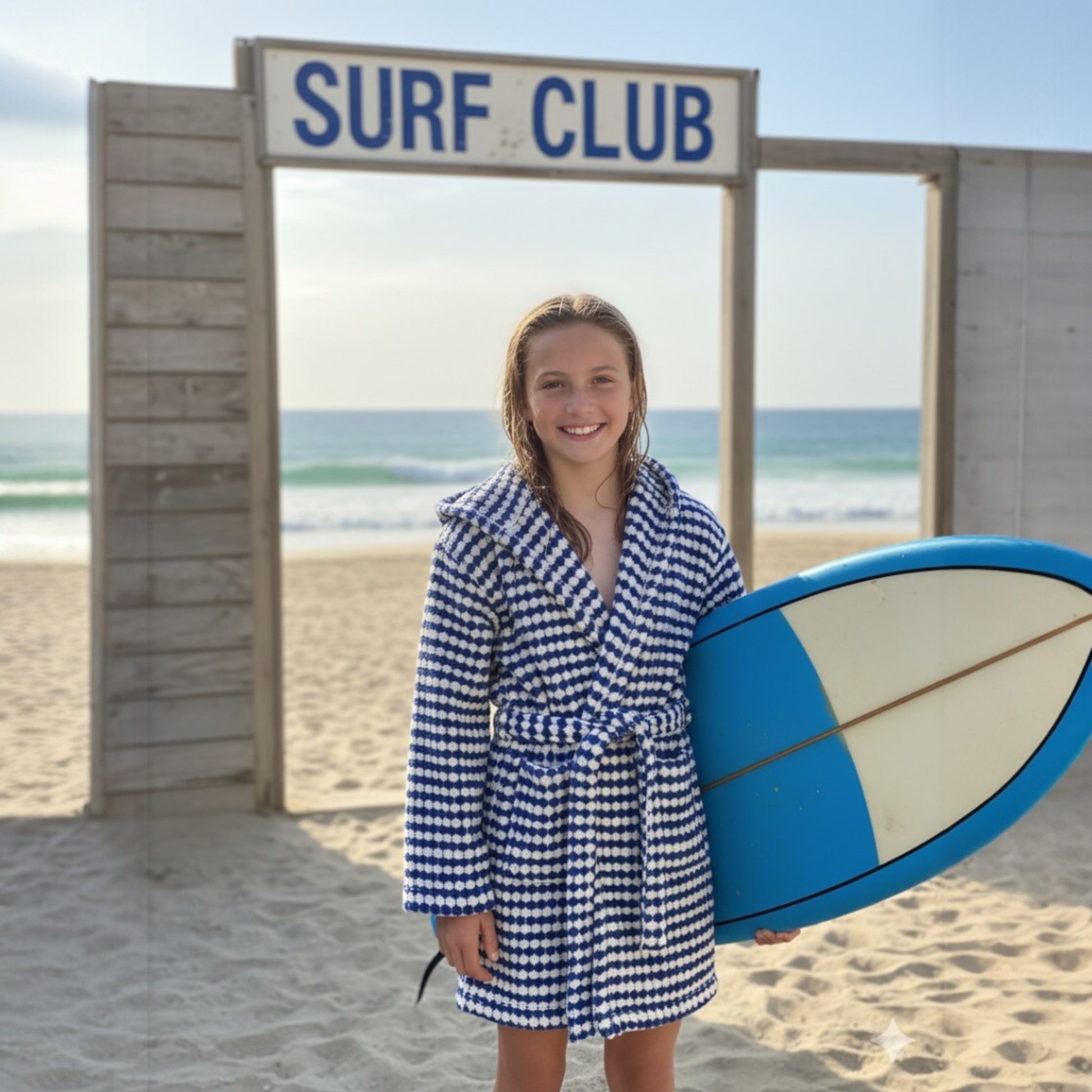 Woman in a blue and white checkered dress holding a blue surfboard in front of a 'Surf Club' sign on a beach.