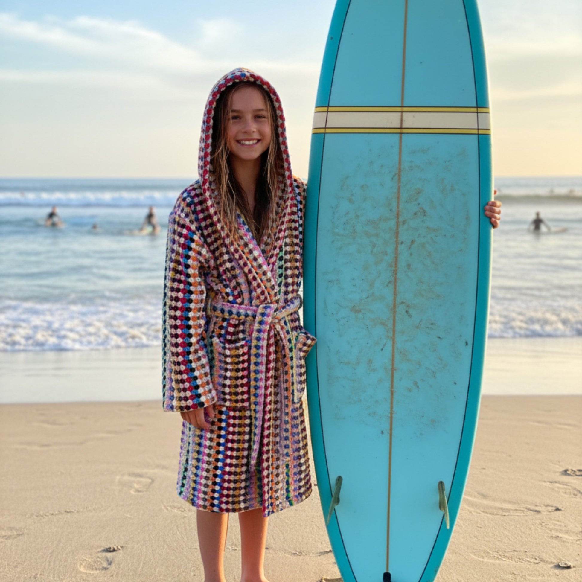 Person in a colorful hooded robe holding a blue surfboard on a beach.