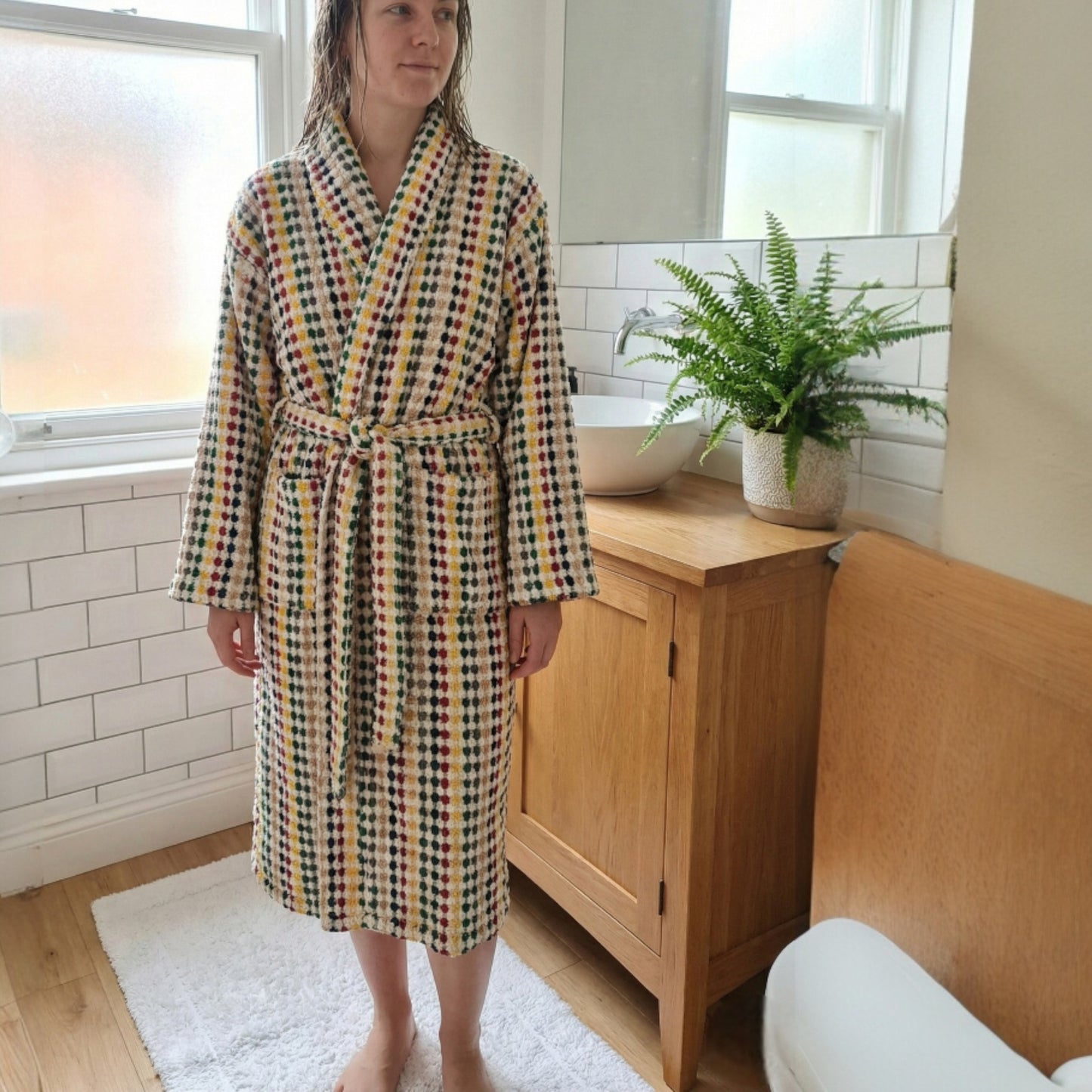 Person wearing a Natural Cotton Hand-Woven Unisex One Size Terry Towel Bathrobe L/XL in a bathroom with a wooden cabinet and plant.